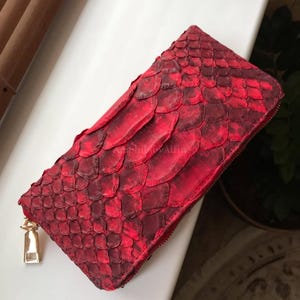 Peut inclure: Un portefeuille rectangulaire pr&eacute;sentant une texture &eacute;cailleuse rouge vif et cramoisi fonc&eacute; sur toute sa surface. Le portefeuille comprend une tirette de fermeture &eacute;clair en m&eacute;tal dor&eacute; et porte l'inscription AmphibbyAlfin sur le devant.