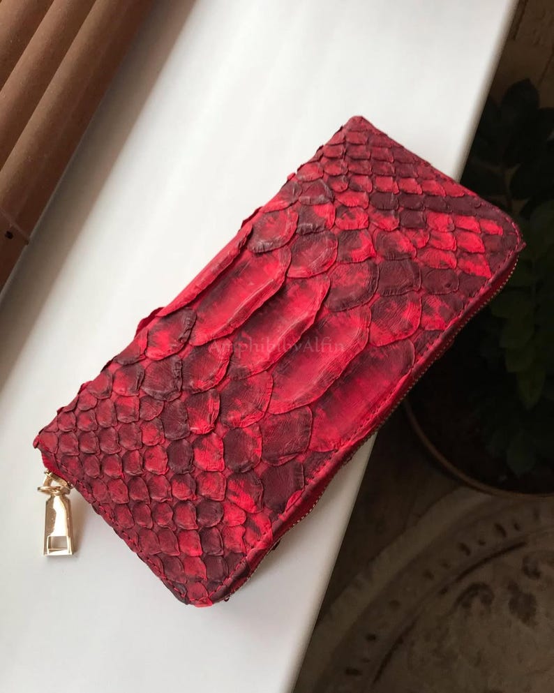 Peut inclure: Un portefeuille rectangulaire pr&eacute;sentant une texture &eacute;cailleuse rouge vif et cramoisi fonc&eacute; sur toute sa surface. Le portefeuille comprend une tirette de fermeture &eacute;clair en m&eacute;tal dor&eacute; et porte l'inscription AmphibbyAlfin sur le devant.