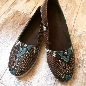 May include: A pair of slip-on flats featuring a textured snakeskin pattern in shades of brown, white, and teal. The shoes have a rounded toe, a dark brown interior lining, and a light-coloured woven jute trim along the base of the sole.