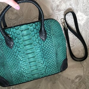 May include: A teal handbag with a textured snakeskin pattern, featuring black leather handles, black corner accents, and a detachable black shoulder strap with silver-toned metal hardware. The bag has a top zip closure and a structured dome shape.
