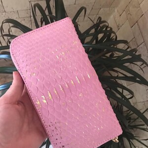 May include: A rectangular pink wallet featuring a textured snakeskin pattern with metallic gold foil accents scattered across the surface. The wallet has a full-length zip closure along the edge and a consistent scale-like embossed finish.