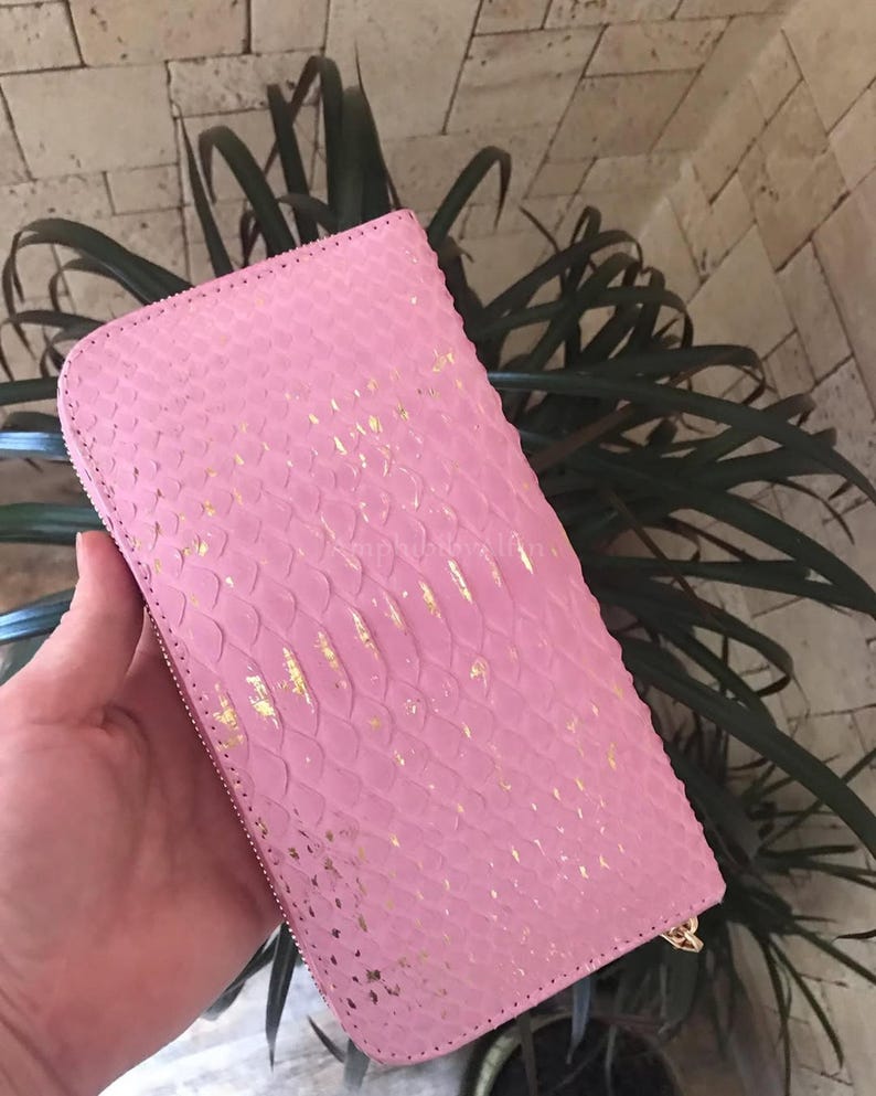 May include: A rectangular pink wallet featuring a textured snakeskin pattern with metallic gold foil accents scattered across the surface. The wallet has a full-length zip closure along the edge and a consistent scale-like embossed finish.