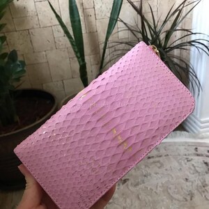 May include: A rectangular wallet featuring a textured pink snakeskin pattern with subtle gold-toned flecks throughout the surface. The wallet has a rounded corner design and a gold-coloured metal zipper closure along the top edge for secure storage.