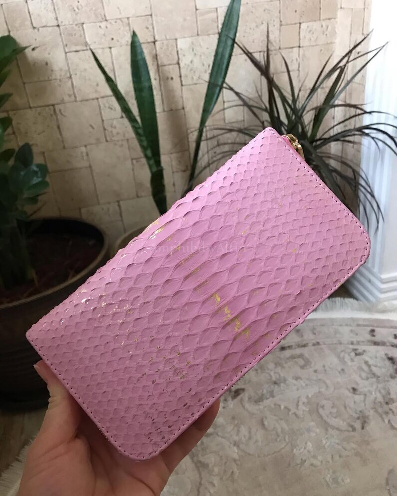May include: A rectangular wallet featuring a textured pink snakeskin pattern with subtle gold-toned flecks throughout the surface. The wallet has a rounded corner design and a gold-coloured metal zipper closure along the top edge for secure storage.