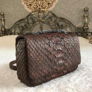 May include: A rectangular brown handbag featuring a textured exterior with overlapping scale-like patterns. The bag includes a metallic silver chain handle attached to the top flap and a matching brown strap extending from the side.