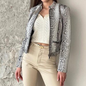 May include: A long-sleeve jacket featuring a grey and white snakeskin pattern with a front zip closure. Underneath, a cream-coloured sleeveless top with a textured chevron knit pattern is paired with high-waisted beige trousers and a belt.