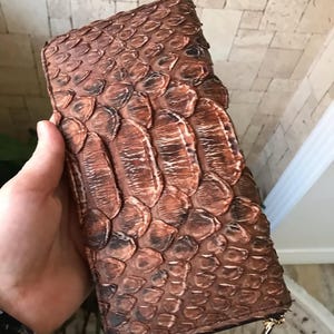 Peut inclure: Un portefeuille rectangulaire présentant une texture en forme d'écailles de couleur brune sur toute sa surface. Le portefeuille est doté d'une fermeture éclair avec une tirette métallique dorée sur le côté, offrant un aspect détaillé et organique.