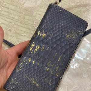 May include: A rectangular crossbody bag featuring a dark gray snakeskin-textured surface with metallic gold accents. The bag includes a thin matching strap and a silver-toned zipper closure at the top, showcasing a distinct scale-like pattern.