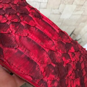 Peut inclure: Un portefeuille rouge pr&eacute;sentant un motif textur&eacute; d'&eacute;cailles superpos&eacute;es sur toute sa surface. Le portefeuille comprend une fermeture &eacute;clair m&eacute;tallique visible sur le bord et affiche le texte AmphibibyAlthie discr&egrave;tement gaufr&eacute;.