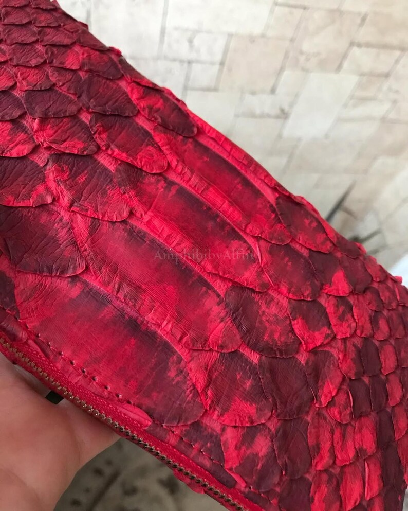 Peut inclure: Un portefeuille rouge pr&eacute;sentant un motif textur&eacute; d'&eacute;cailles superpos&eacute;es sur toute sa surface. Le portefeuille comprend une fermeture &eacute;clair m&eacute;tallique visible sur le bord et affiche le texte AmphibibyAlthie discr&egrave;tement gaufr&eacute;.