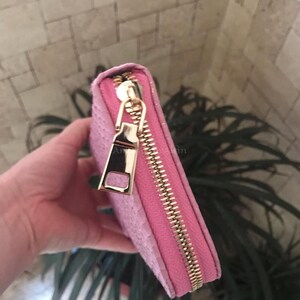 May include: A pink wallet with a textured, snakeskin-like pattern on the exterior. The wallet features a gold-toned metal zip that runs along the edge, complete with a matching gold-toned rectangular zip pull tab for easy opening and closing.