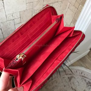 Peut inclure: Un portefeuille rouge avec un ext&eacute;rieur textur&eacute; et une fermeture &eacute;clair dor&eacute;e. L'int&eacute;rieur est doubl&eacute; de tissu rouge et comprend plusieurs fentes pour cartes, un compartiment central zipp&eacute; et des sections ouvertes pour organiser les billets.