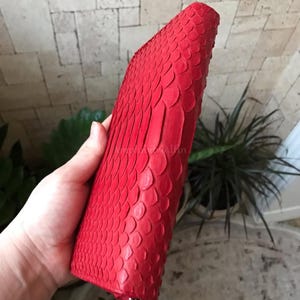 Peut inclure: Un portefeuille rouge vif pr&eacute;sentant un motif textur&eacute; en forme d'&eacute;cailles sur toute sa surface ext&eacute;rieure. Le portefeuille a une forme rectangulaire avec des bords arrondis et une tirette de fermeture &eacute;clair en m&eacute;tal argent&eacute; visible en bas.