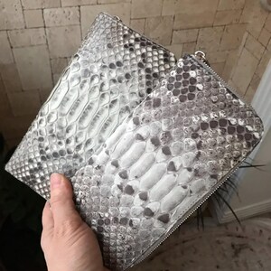 Op de afbeelding: Twee rechthoekige portemonnees met een gestructureerd slangenleerpatroon in grijs en wit. Elke portemonnee heeft een zilverkleurige metalen ritssluiting langs de boven- en zijkanten, met een consistente schubachtige textuur over het oppervlak.