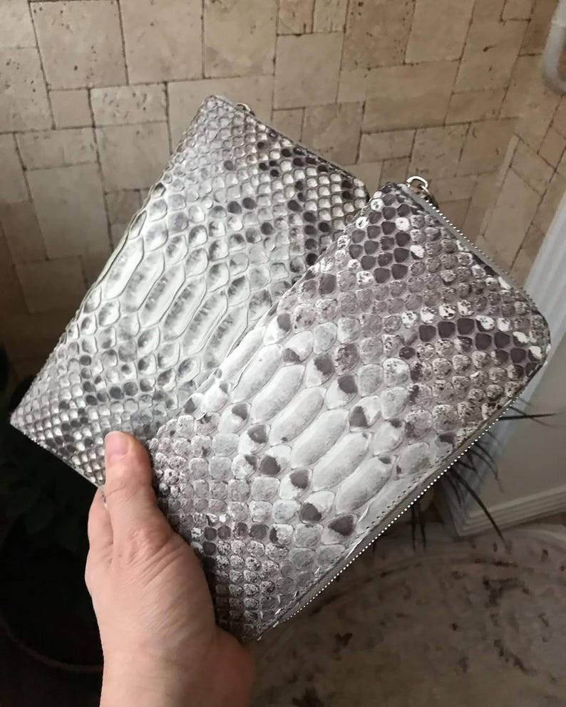 Op de afbeelding: Twee rechthoekige portemonnees met een gestructureerd slangenleerpatroon in grijs en wit. Elke portemonnee heeft een zilverkleurige metalen ritssluiting langs de boven- en zijkanten, met een consistente schubachtige textuur over het oppervlak.