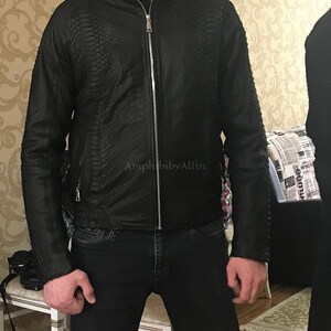 May include: A black jacket with a textured snakeskin-like pattern, featuring a full-length silver-tone metal zip closure, a stand-up collar, and long sleeves. The text AmphibibyAlfin is visible on the front of the black jacket.