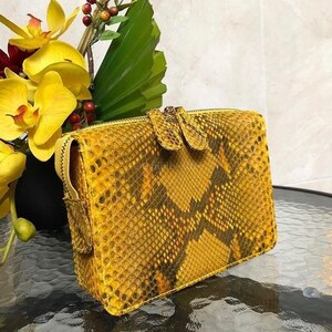 Könnte beinhalten: Eine rechteckige Clutch mit einem leuchtend gelben und dunkelbraunen Schlangenledermuster. Die Tasche verfügt über einen goldfarbenen Reißverschluss mit zwei passenden Schlangenleder-Reißverschlussanhängern für ein sicheres Design.