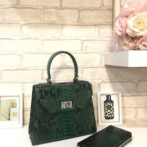 May include: A dark green snakeskin-textured handbag with a top handle and a silver-toned metal turn-lock closure, accompanied by a matching rectangular zippered wallet with a silver-toned pull tab, both featuring a distinct reptilian scale pattern.