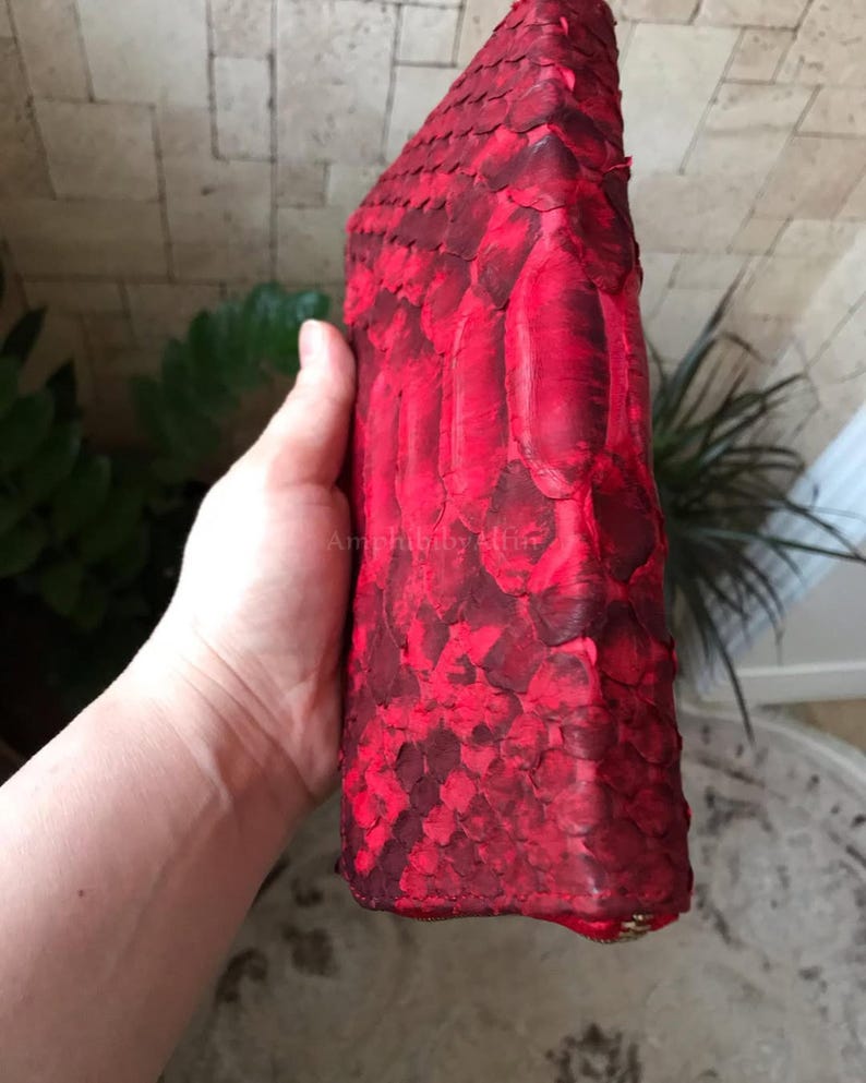 Peut inclure: Un portefeuille rouge vif pr&eacute;sentant une surface textur&eacute;e semblable &agrave; des &eacute;cailles. Le portefeuille a une forme rectangulaire avec des bords arrondis et une fermeture &eacute;clair visible en bas. La surface affiche des nuances de rouge vari&eacute;es.