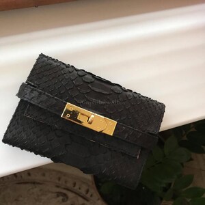 May include: A black wallet with a textured snakeskin pattern featuring a horizontal strap across the front. The wallet is secured with a polished gold-tone metal clasp closure. The text AmphibibyAlfin is faintly visible on the center of the wallet.