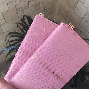 May include: A rectangular pink wallet featuring a textured snakeskin-like pattern with gold-coloured foil accents. The wallet has a gold-coloured metal zip closure along the top edge and a smooth pink interior lining visible at the opening.