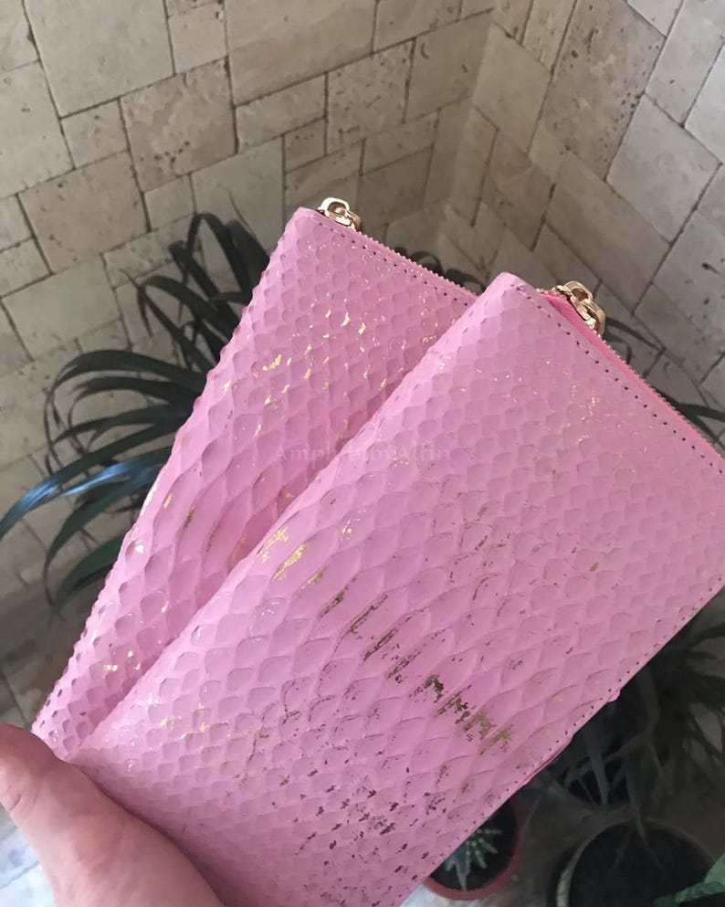 May include: A rectangular pink wallet featuring a textured snakeskin-like pattern with gold-coloured foil accents. The wallet has a gold-coloured metal zip closure along the top edge and a smooth pink interior lining visible at the opening.