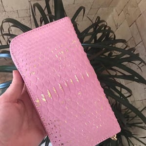 May include: A rectangular zip-around wallet featuring a textured pink surface with a repeating scale-like pattern and irregular gold-coloured foil accents scattered across the exterior. The wallet has a matching pink zipper along the edge.