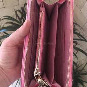 May include: A bright pink zip-around wallet with a textured exterior and a matching pink interior. The wallet features a central zippered coin compartment, multiple card slots, and gold-toned metal hardware. The text AmibibyAlfin is visible.