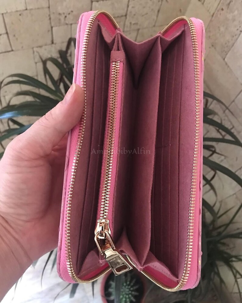 May include: A bright pink zip-around wallet with a textured exterior and a matching pink interior. The wallet features a central zippered coin compartment, multiple card slots, and gold-toned metal hardware. The text AmibibyAlfin is visible.
