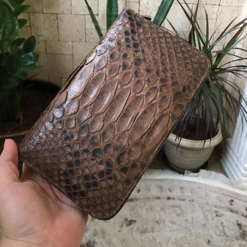 May include: A rectangular clutch wallet featuring a textured brown snakeskin pattern with overlapping scales. The wallet has a zip-around closure along the edge and a consistent, natural gradient of dark and light brown tones across the surface.