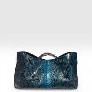 May include: A teal and black snakeskin patterned handbag with a structured rectangular body and a curved, metallic-toned handle. The bag features a distinct scale texture throughout the exterior surface, creating a dark, multi-tonal appearance.