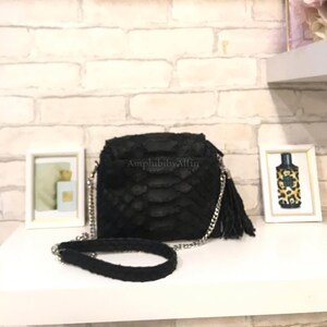 Peut inclure: Un sac bandoulière noir texturé avec un motif imitation peau de serpent, doté d'une chaîne argentée, d'une bandoulière tressée noire et d'un pompon latéral. Le sac porte l'inscription AmphibibyAlfin sur le rabat avant et possède des détails argentés.