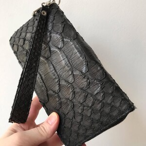 May include: A dark charcoal grey rectangular wristlet wallet featuring a textured, overlapping scale pattern across the entire exterior surface. The wallet includes a matching wrist strap attached to a silver-toned metal zipper pull at the top corner.
