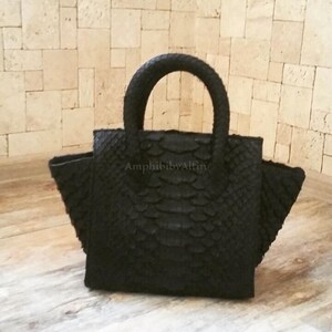 May include: A black handbag with a textured, scale-like surface pattern and a rounded top handle. The bag features a structured, trapezoidal silhouette with flared sides and the text AmphibibyAlfin printed in the centre of the front panel.