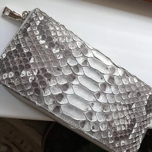 Op de afbeelding: Een rechthoekige portemonnee met een gestructureerd grijs en wit slangenleerpatroon. De portemonnee heeft een zilverkleurige metalen ritssluiting langs de bovenrand, met een bijpassende zilverkleurige metalen ritstrekker aan de zijkant.