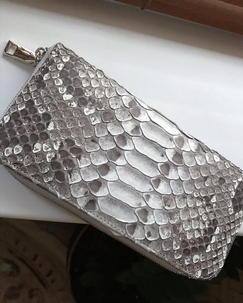 Op de afbeelding: Een rechthoekige portemonnee met een gestructureerd grijs en wit slangenleerpatroon. De portemonnee heeft een zilverkleurige metalen ritssluiting langs de bovenrand, met een bijpassende zilverkleurige metalen ritstrekker aan de zijkant.