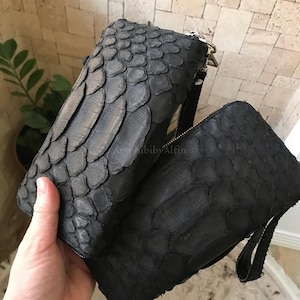 May include: Two rectangular black wristlet pouches featuring a textured, scale-like pattern across the exterior surface. Each pouch includes a secure zipper closure along the top edge and a matching black wrist strap attached to the side hardware.