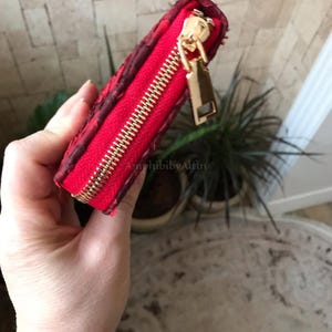 Peut inclure: Un petit portefeuille rectangulaire rouge avec une surface textur&eacute;e et une fermeture &eacute;clair en m&eacute;tal dor&eacute;. La tirette de la fermeture &eacute;clair est une languette rectangulaire en m&eacute;tal dor&eacute;. Le portefeuille pr&eacute;sente une doublure rouge.