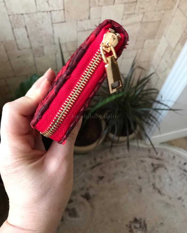 Peut inclure: Un petit portefeuille rectangulaire rouge avec une surface textur&eacute;e et une fermeture &eacute;clair en m&eacute;tal dor&eacute;. La tirette de la fermeture &eacute;clair est une languette rectangulaire en m&eacute;tal dor&eacute;. Le portefeuille pr&eacute;sente une doublure rouge.