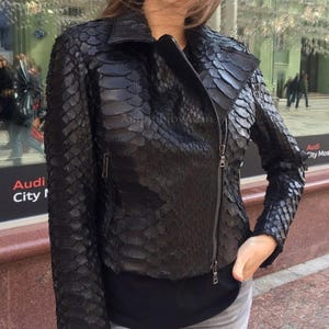May include: A black jacket featuring a textured, scale-like pattern across the entire surface. The jacket has a collared neckline, long sleeves, and a diagonal metal zip closure extending from the waist to the upper chest area.
