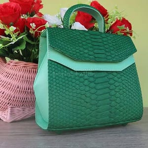 May include: A structured handbag featuring a vibrant green snakeskin-textured flap and body, accented by smooth light mint green side panels and a matching top handle. The bag has a geometric silhouette with a fold-over closure and a clean design.