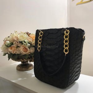 Peut inclure: Un sac seau noir avec un motif texturé en peau de serpent et des anses en chaîne dorée. Le sac présente une silhouette structurée et est placé à côté d'un bol décoratif rempli de roses artificielles de couleur crème et pêche.
