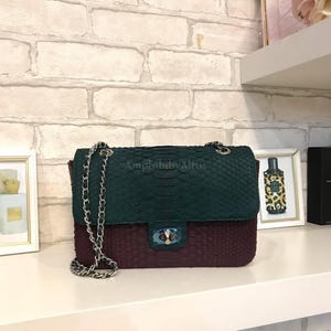 May include: A rectangular shoulder bag featuring a two-tone snakeskin texture with a dark green upper flap and a deep burgundy lower body. The bag includes a silver-toned chain strap and a central silver-toned twist-lock closure with blue film.