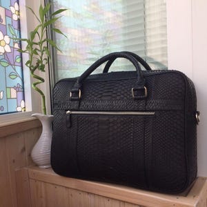May include: A black briefcase with a textured snakeskin pattern, featuring two top handles, a horizontal front zipped pocket with silver-toned hardware, and a rectangular silhouette. The bag has a structured design with visible stitching details.