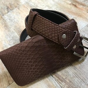 May include: A brown belt with a textured snakeskin pattern and a silver-toned metal buckle. The belt features a loop and a series of holes for adjustment. The text AmphibibyAlfin is visible on the surface of the belt strap near the buckle.