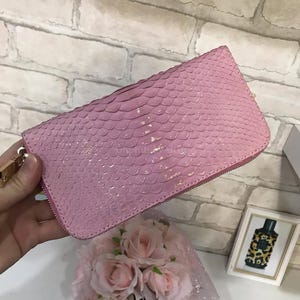 May include: A rectangular wallet with a pink, textured, scale-like pattern across the exterior surface. The wallet features a metallic zip closure along the top and side edges, with a gold-toned metal zip pull attached to the side.