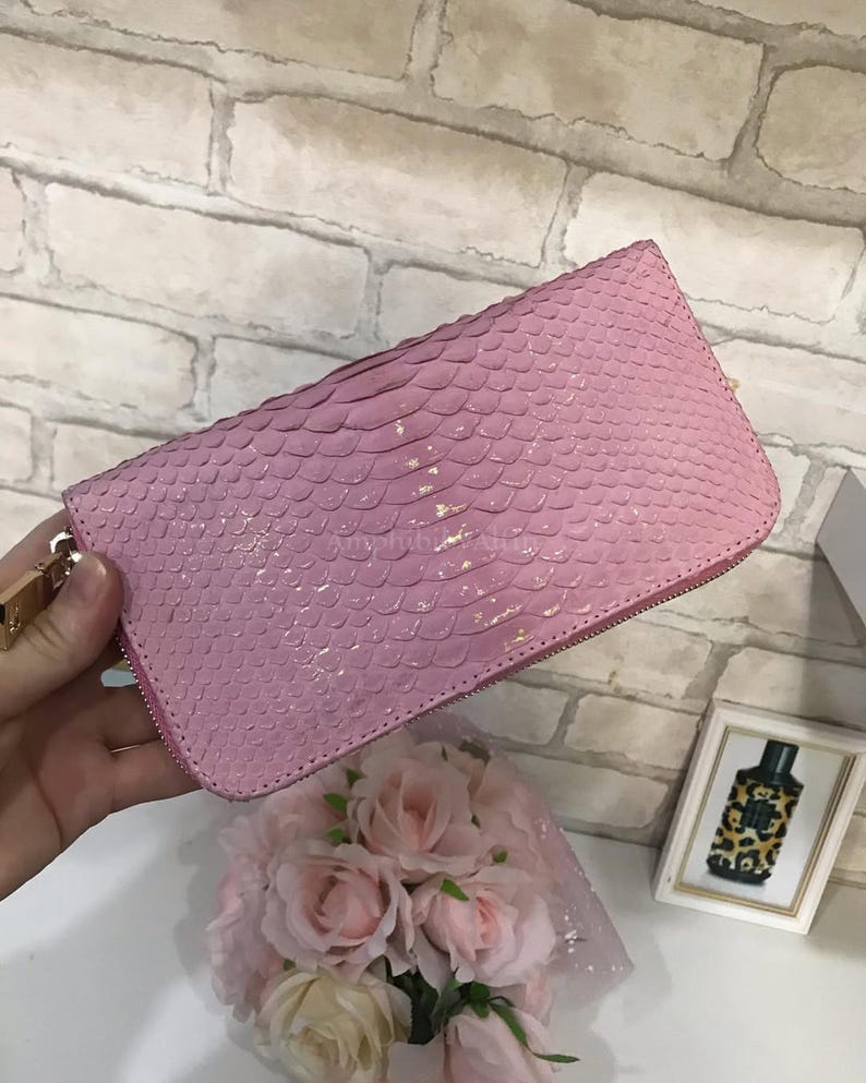 May include: A rectangular wallet with a pink, textured, scale-like pattern across the exterior surface. The wallet features a metallic zip closure along the top and side edges, with a gold-toned metal zip pull attached to the side.