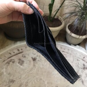 以下が含まれることがあります： 質感のある外装表面と複数のカードスロットを備えた黒い二つ折り財布。内側の裏地は黒で、財布の内側の折り目にはAmphibibyAlfiというブランド名が小さな明るい色の文字で印刷されています。