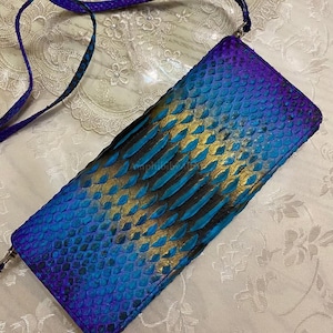 May include: A rectangular crossbody bag featuring a textured snakeskin pattern with a gradient color scheme transitioning from deep purple to vibrant blue and metallic gold. The bag includes a matching thin shoulder strap and a metal zipper closure.