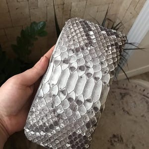 May include: A rectangular wallet featuring a textured snakeskin pattern in shades of grey, white, and black. The wallet has a zip-around closure with a silver-toned metal zipper pull, showcasing a detailed scale design across the entire surface.